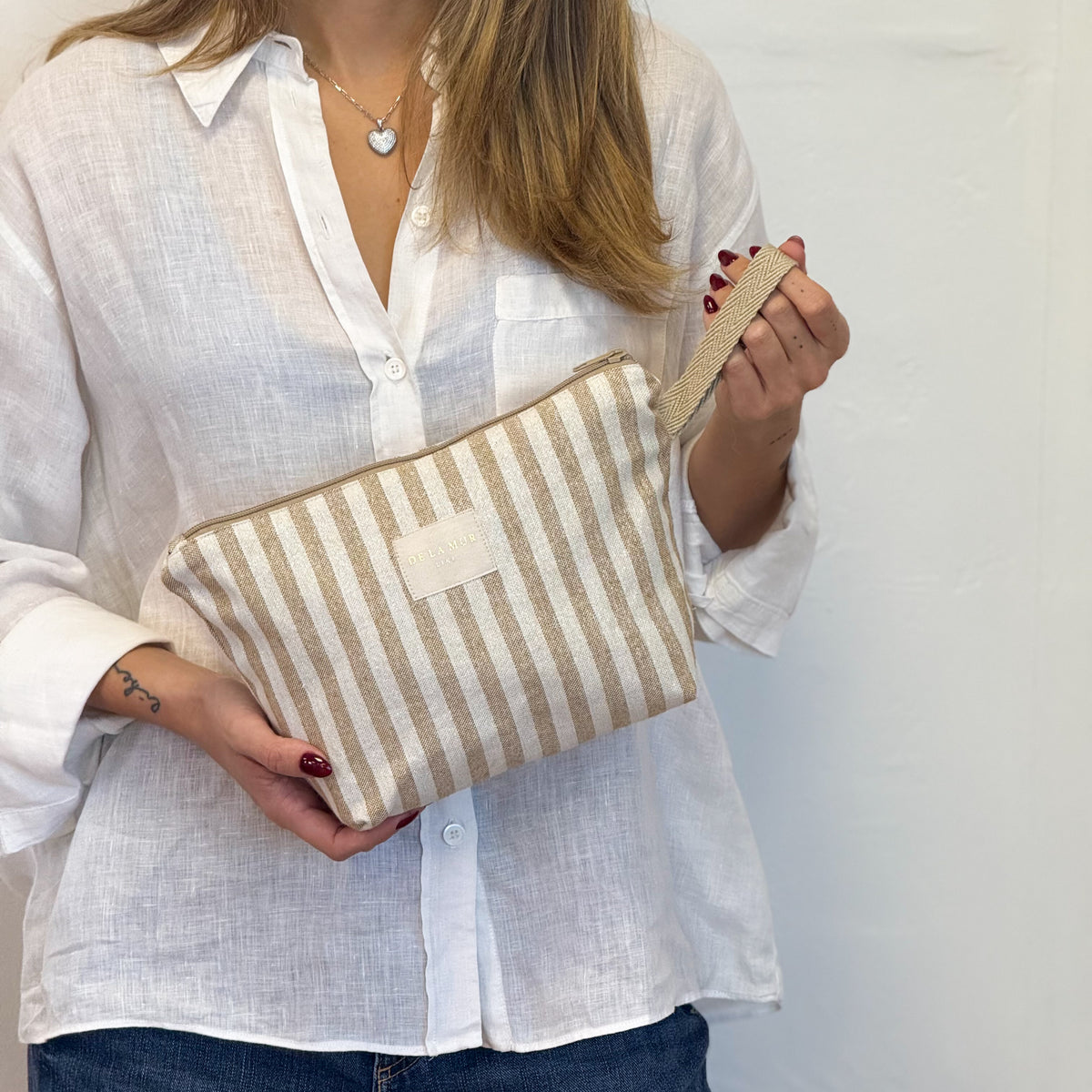 Model holding the beige striped DE LA MUR Lumos makeup bag by its wrist strap